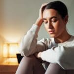 Woman sitting indoors with eyes closed and head resting on hand, appearing overwhelmed or emotionally drained
