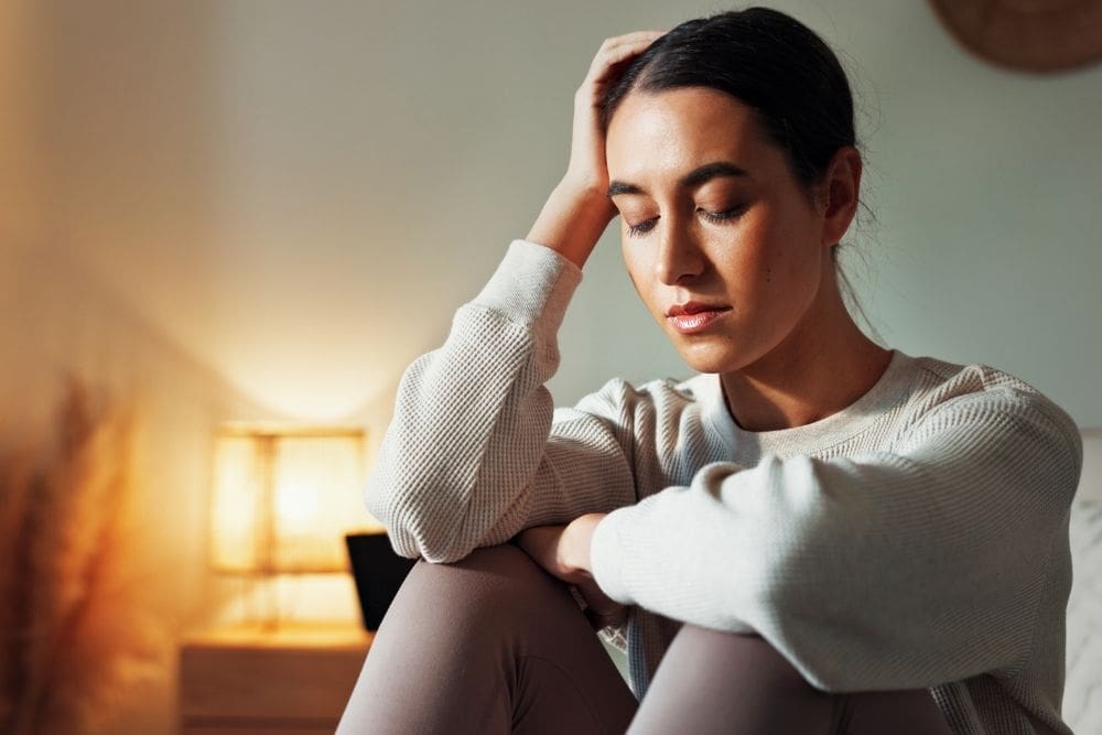 Woman sitting indoors with eyes closed and head resting on hand, appearing overwhelmed or emotionally drained