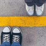 Two pairs of sneakers facing each other on opposite sides of a bold yellow line painted on concrete.