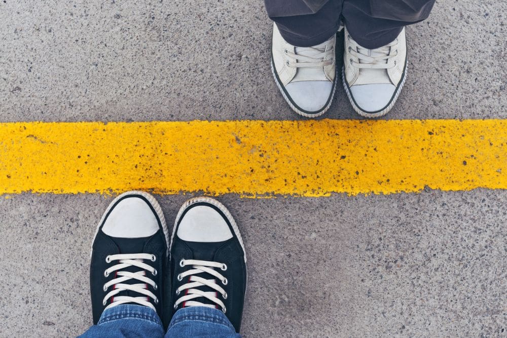 Two pairs of sneakers facing each other on opposite sides of a bold yellow line painted on concrete.