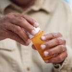 Close-up of a person’s hands opening an orange prescription pill bottle, wearing a beige shirt.