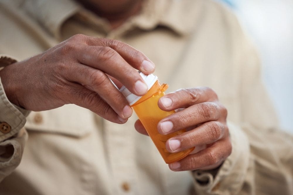 Close-up of a person’s hands opening an orange prescription pill bottle, wearing a beige shirt.