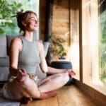Young woman meditating cross-legged indoors by a large window, smiling peacefully