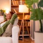 A woman relaxing on a white sofa with her hands behind her head, smiling peacefully in a cozy living room with warm lighting, bookshelves, and houseplants.