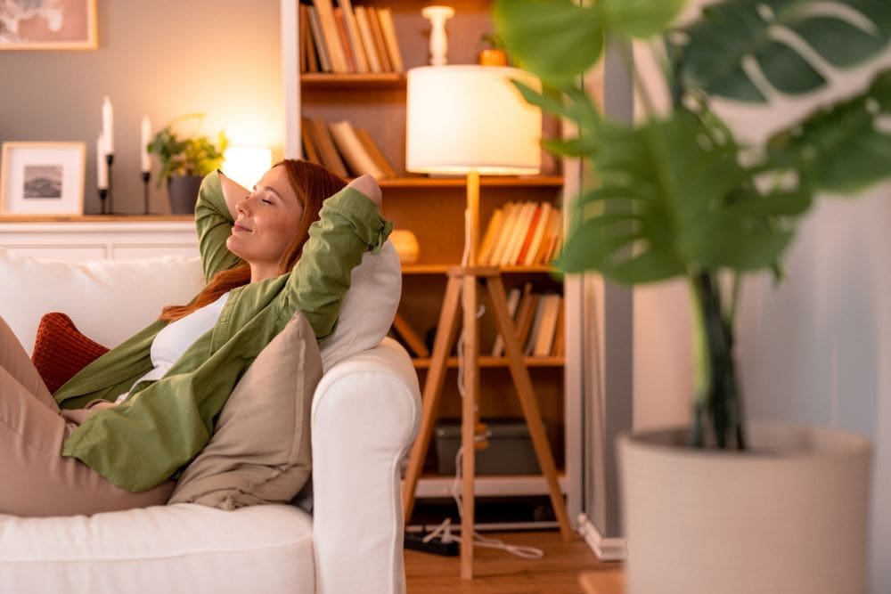 A woman relaxing on a white sofa with her hands behind her head, smiling peacefully in a cozy living room with warm lighting, bookshelves, and houseplants.