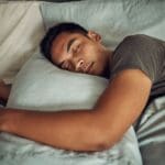 A young man peacefully sleeping on his side in bed, hugging a pillow, with soft natural light coming through a nearby window.