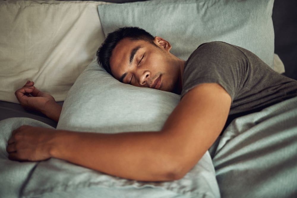 A young man peacefully sleeping on his side in bed, hugging a pillow, with soft natural light coming through a nearby window.
