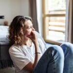 A woman sits on the floor by her bed with her knees drawn up, resting her hand on her chin. She looks out a bright window with a thoughtful, worried expression.