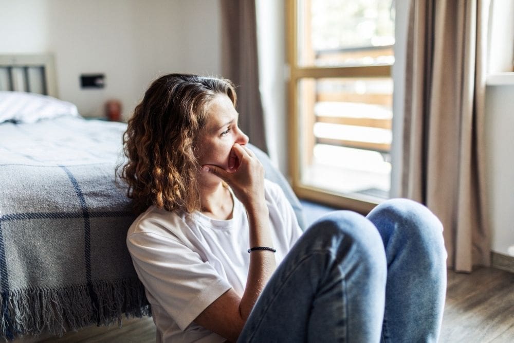 A woman sits on the floor by her bed with her knees drawn up, resting her hand on her chin. She looks out a bright window with a thoughtful, worried expression.
