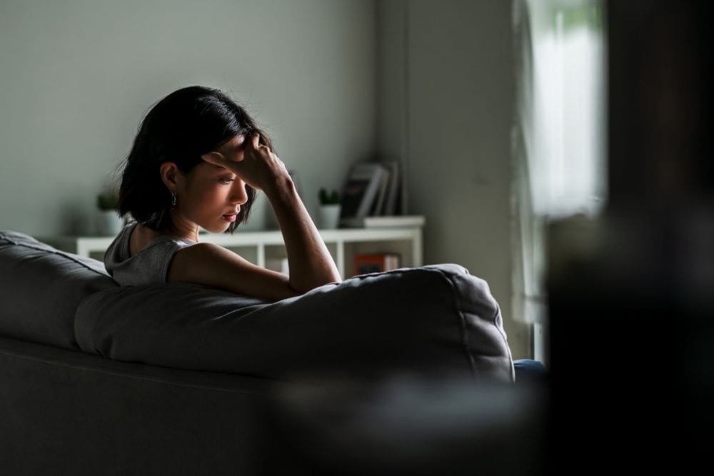 Woman sitting on a couch in a dimly lit room, resting her head in her hand with a pensive expression, suggesting stress or emotional fatigue.