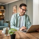 A smiling man wearing glasses uses a laptop at a wooden table in a cozy living room, with a plant and glass of water in front of him.