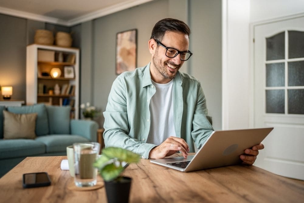 A smiling man wearing glasses uses a laptop at a wooden table in a cozy living room, with a plant and glass of water in front of him.
