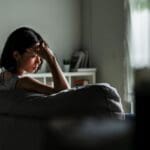 Woman sitting on a couch in a dimly lit room with her hand on her forehead, appearing deep in thought or emotionally distressed.