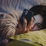 Young woman lying in bed at night looking at her smartphone with a tired expression.