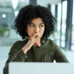 A woman sits at a desk looking thoughtful and anxious, resting her hand near her mouth as she stares ahead in a bright office setting.