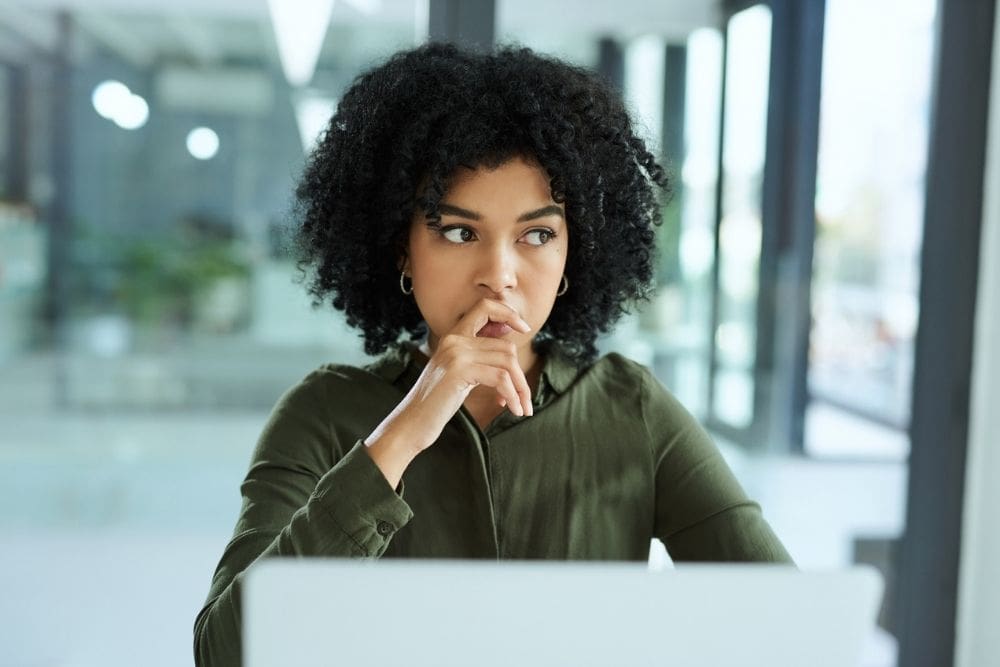 A woman sits at a desk looking thoughtful and anxious, resting her hand near her mouth as she stares ahead in a bright office setting.