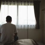 Man sitting on the edge of a bed facing a window, appearing deep in thought in a dimly lit room.