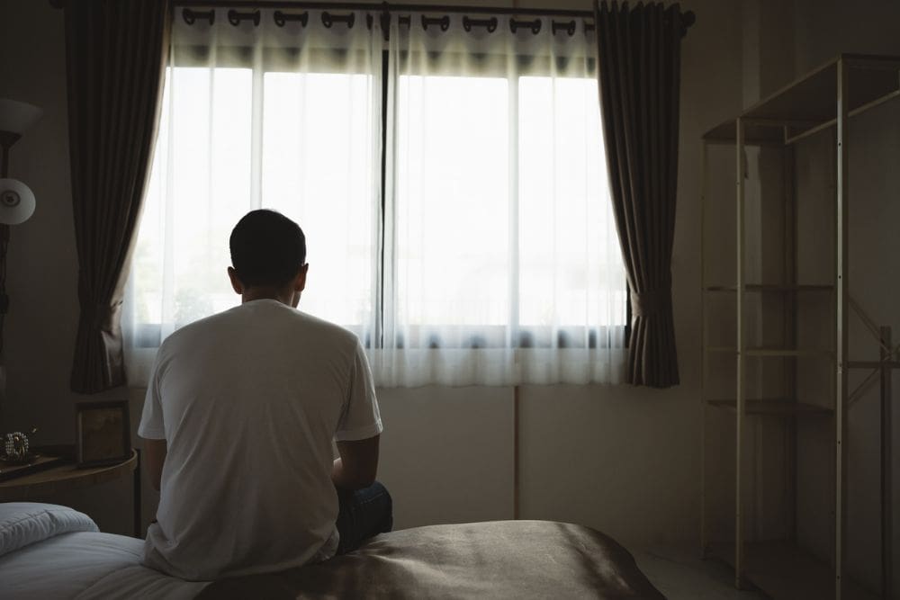 Man sitting on the edge of a bed facing a window, appearing deep in thought in a dimly lit room.
