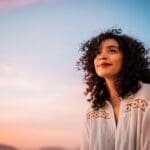 Woman with curly hair smiling softly while looking up at the colorful sunset sky, wearing a white blouse and appearing peaceful and hopeful.