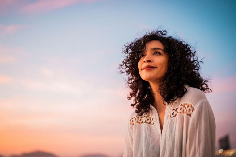 Woman with curly hair smiling softly while looking up at the colorful sunset sky, wearing a white blouse and appearing peaceful and hopeful.