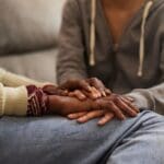 Two people sit close together on a couch, one gently holding the other’s hands in a quiet gesture of comfort and emotional support.