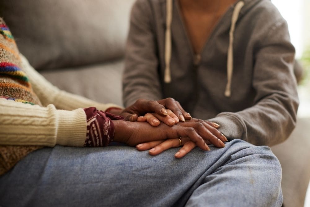 Two people sit close together on a couch, one gently holding the other’s hands in a quiet gesture of comfort and emotional support.