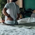 A person sitting on the edge of a bed with one hand on their lower back, appearing to experience back pain or discomfort.