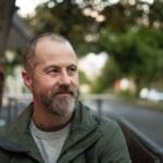 A middle-aged man with a beard stands outdoors on a tree-lined street, looking ahead with a calm, reflective expression.