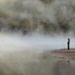 Person standing alone by a fog-covered body of water, looking into the distance, conveying uncertainty and quiet reflection.