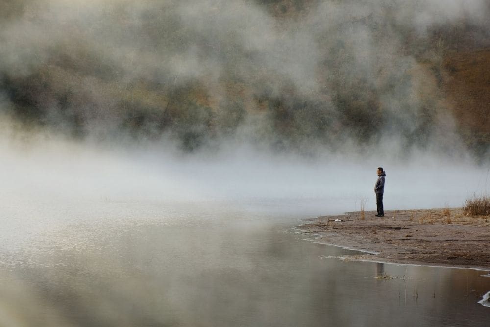 Person standing alone by a fog-covered body of water, looking into the distance, conveying uncertainty and quiet reflection.