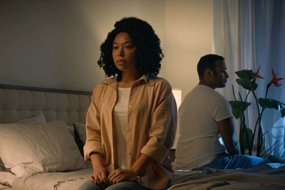 Raleigh Oaks - The Anxiety-Avoidance Cycle Understanding Why Your Anxiety Grows When You Avoid Things. A frustrated couple sits in a dimly lit bedroom. The woman faces away from the man as she looks upset and disgruntled.