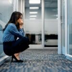 Raleigh Oaks - Coping With Bipolar Disorder at Work. A woman squats down with an axious look in her face in the hall of a professional office building