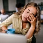 Raleigh Oaks - How Anxiety Affects Sleep - Edited. A woman with a cup of coffee sits at a table rubbing her eyes with exhaustion.